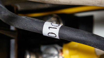 Elektrik kutusunun siyah kablosunun etrafına siyah metinle yazılmış beyaz, kendi kendine sönme özellikli Tedlar sarma etiket.